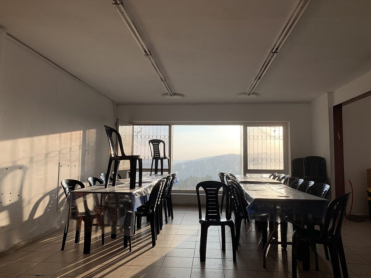 An empty classroom with plastic chairs and tables, lit by daylight from a bank of windows.