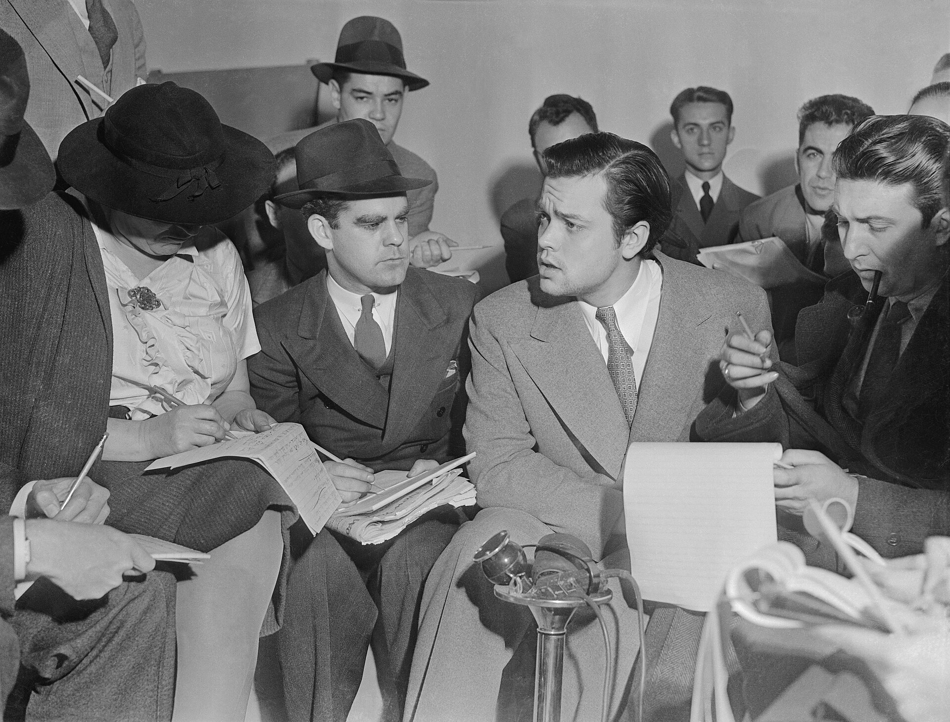 Orson Welles standing with raised arms addressing a group of newspaper reporters the day after the 1938 broadcast.